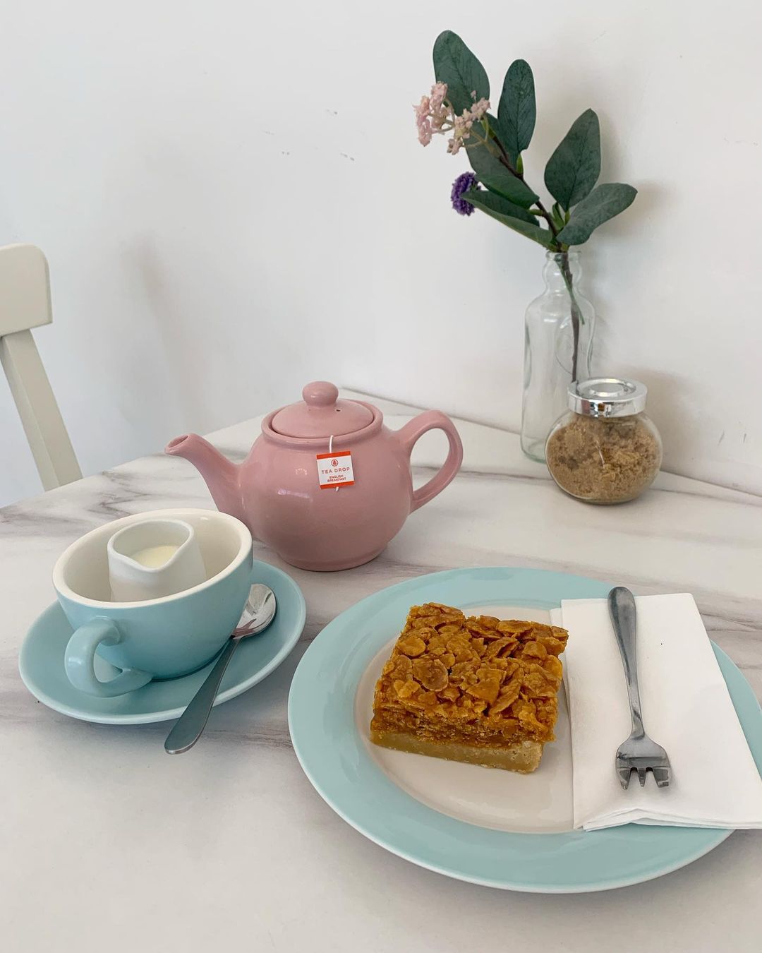 A table, on which sits a pink teapot, a cup of coffee and a slice of cornflake cake.