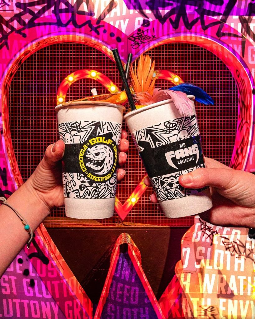 Two people clink glasses in front of a heart background at Golf Fang Liverpool.