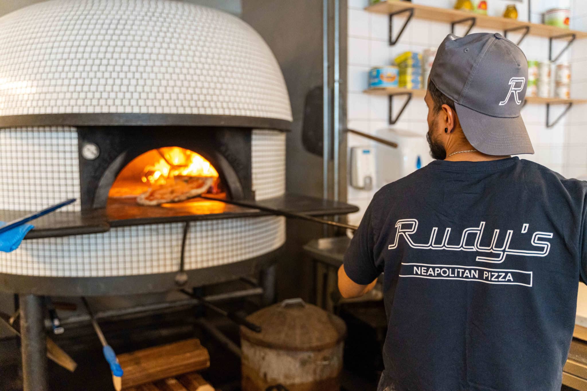A man placing a pizza into a white pizza oven, wearing a tshirt that says 'Rudy's'.