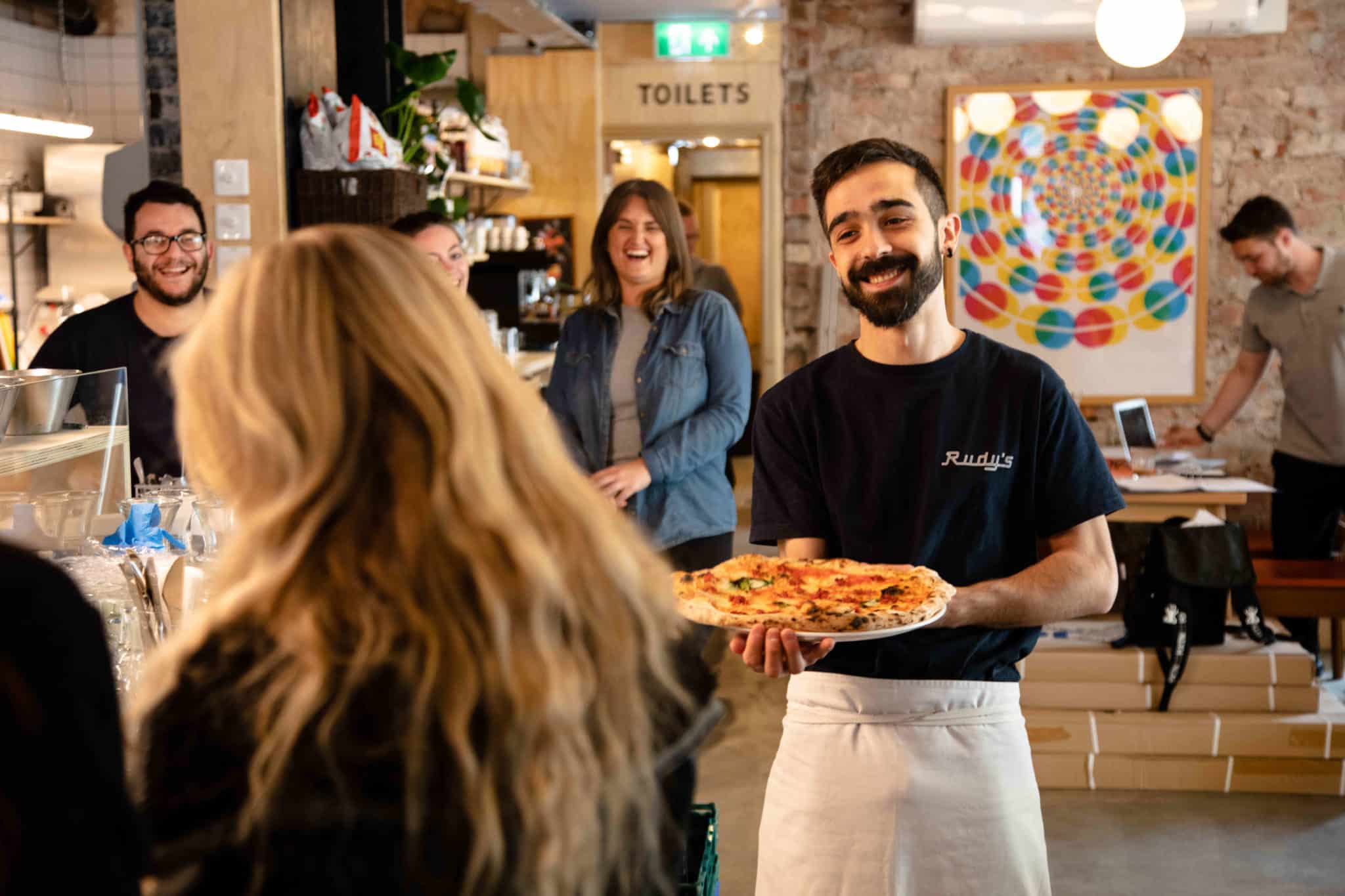 A smiling waiter bringing a pizza to a woman in Rudy's Pizzeria.