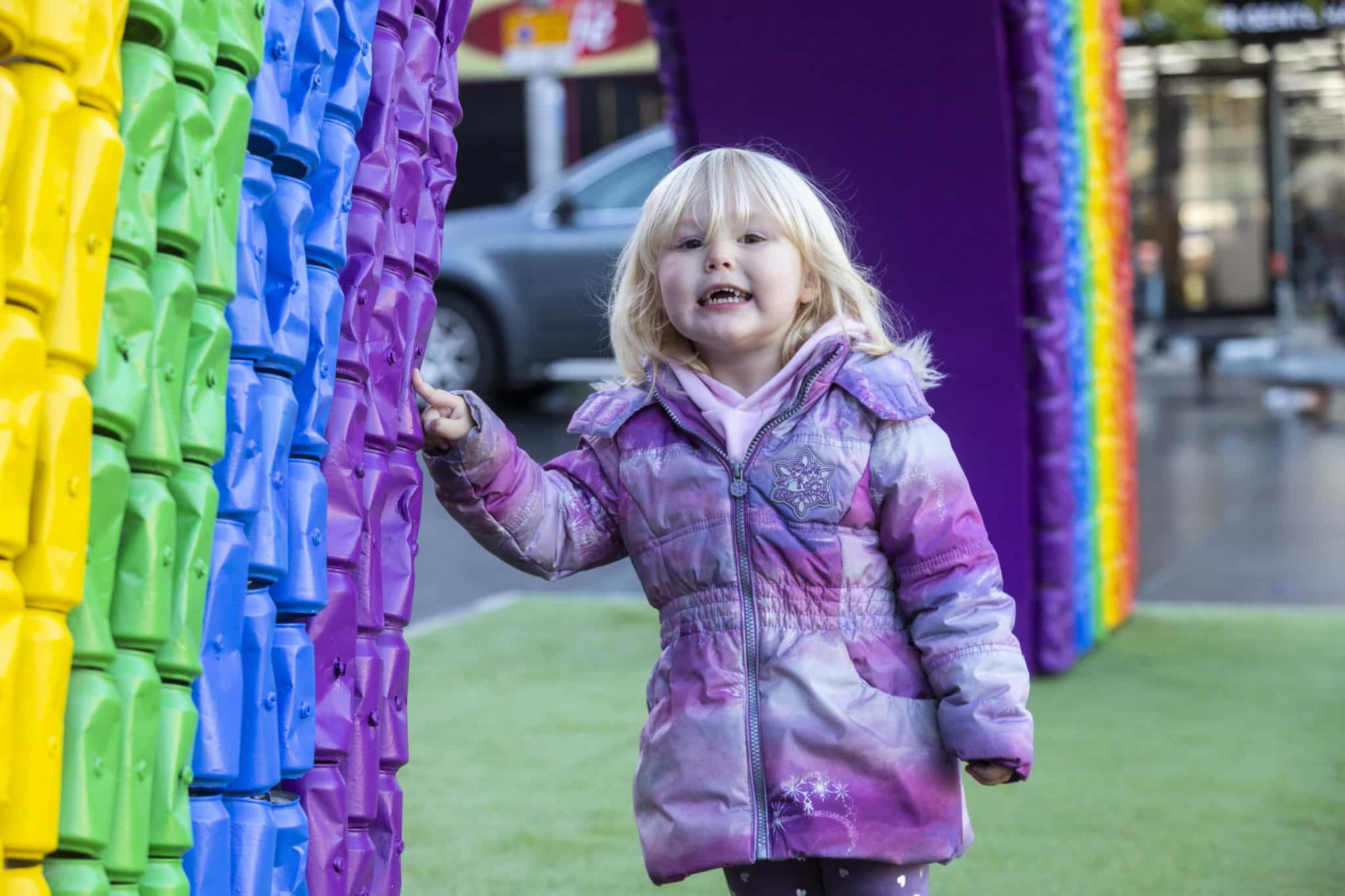 A little girl stands by the giant recycled rainbow in Liverpool city centre, smiling. 