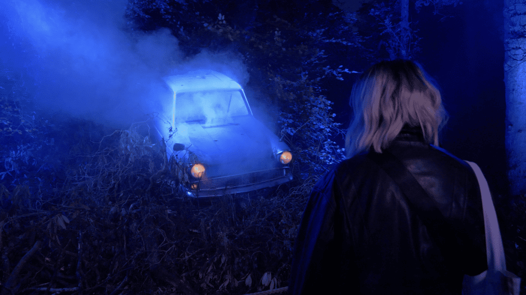 A blue Ford Anglia sits illuminated in a forest, observed by a woman. 