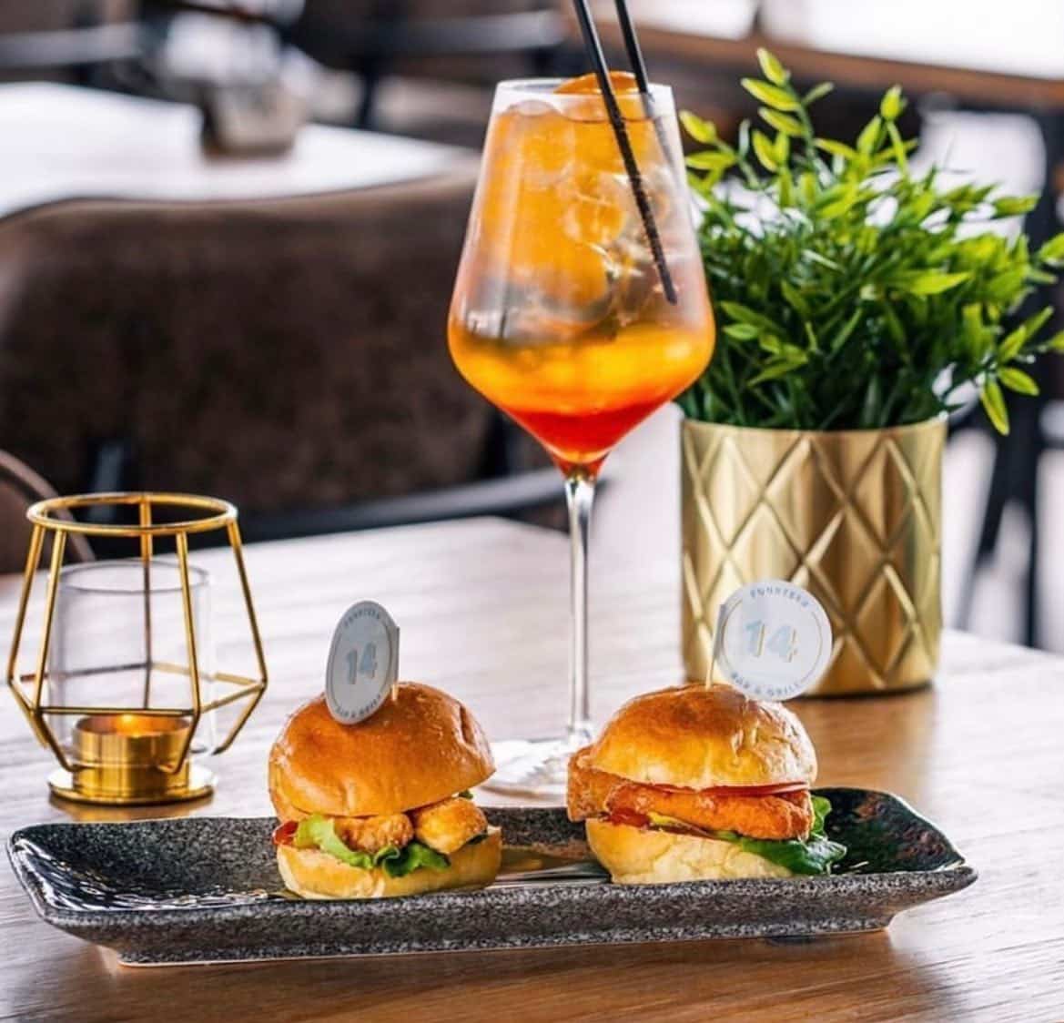 An orange cocktail beside a plate on which are two sliders are 14 Bar & Grill.
