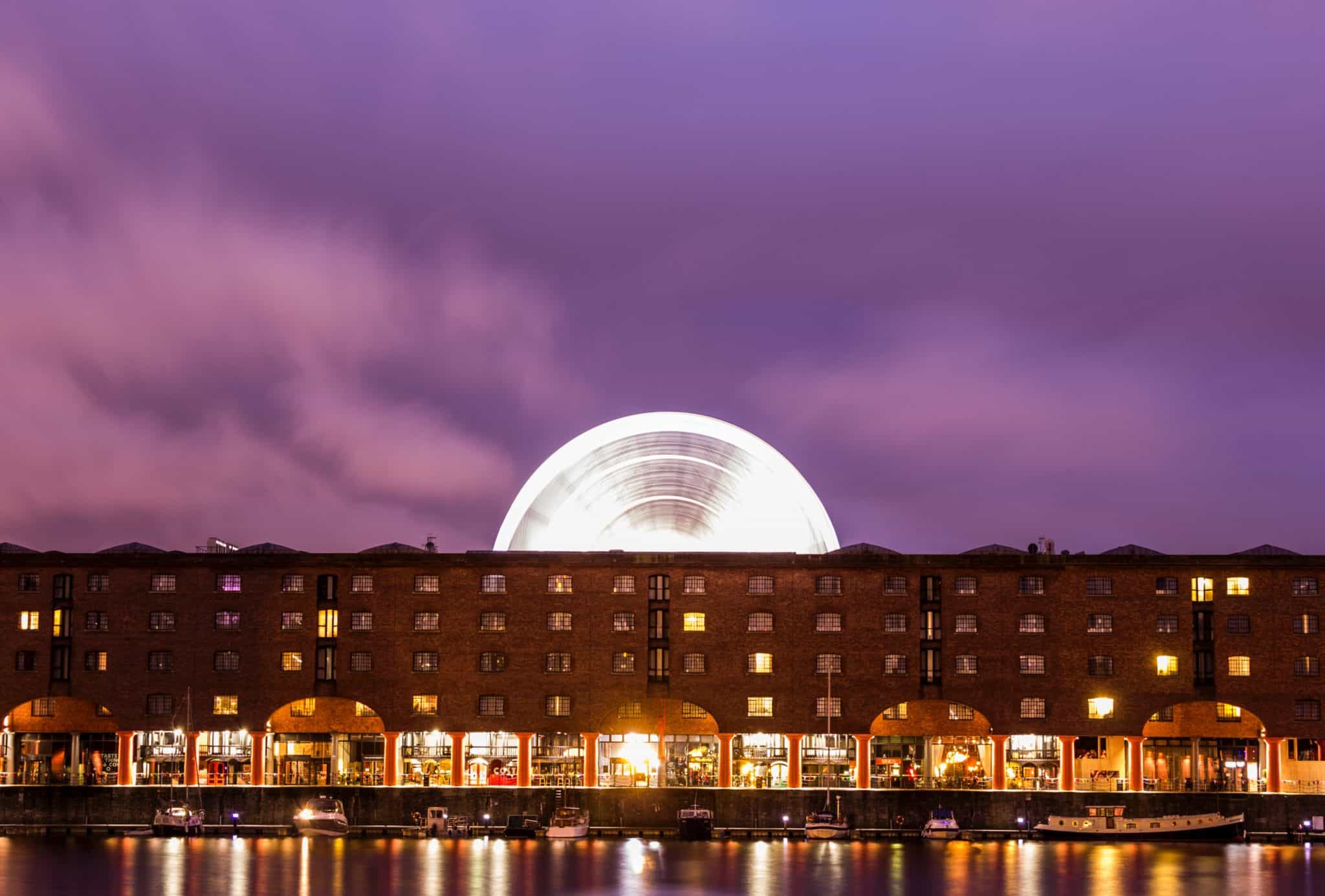 The Tate Liverpool lit up at night with a purple sky and light bouncing of the water. 